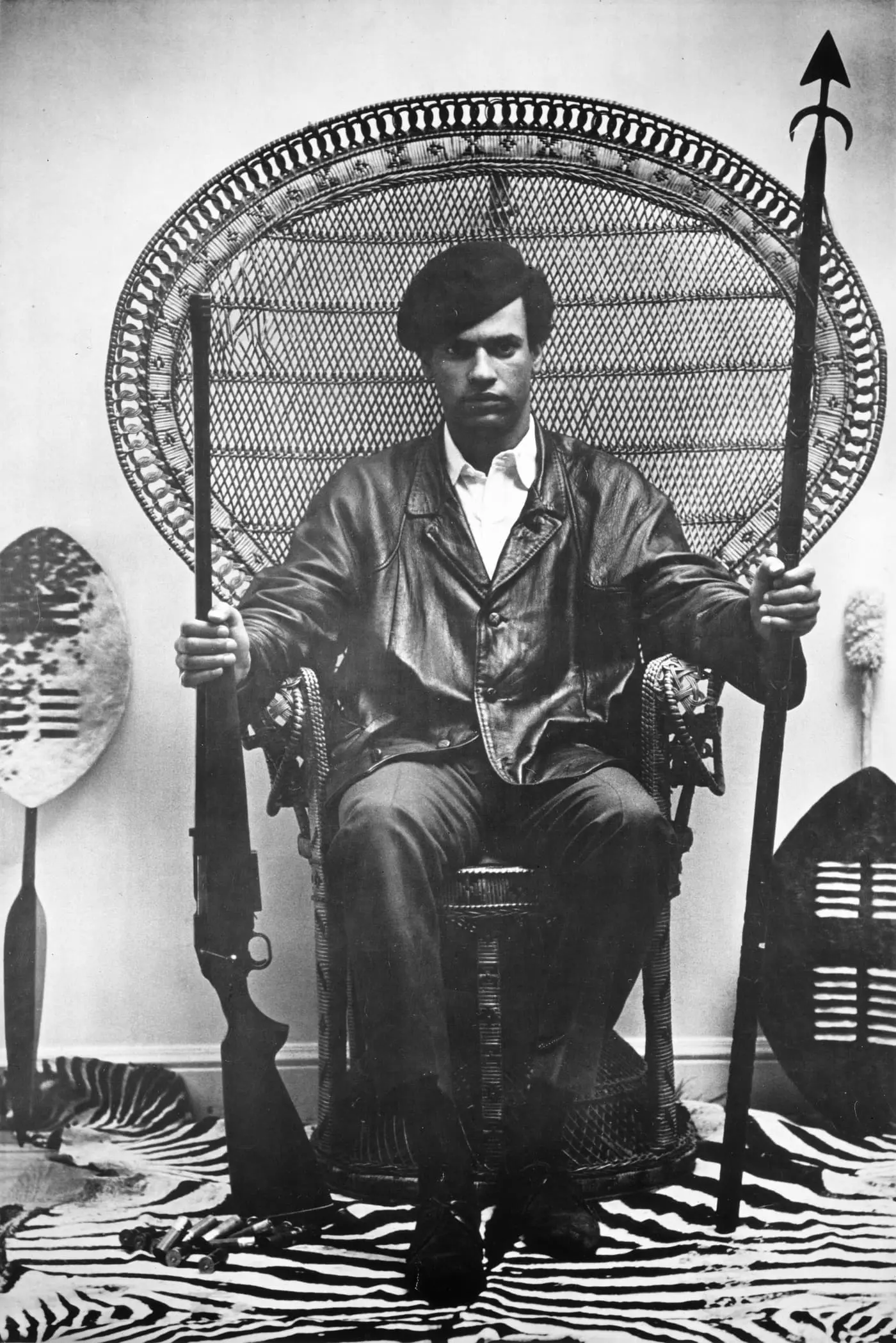 Huey P. Newton, cofondateur du Black Panther Party, assis dans le célèbre fauteuil en rotin, lance et fusil en main, vers 1968